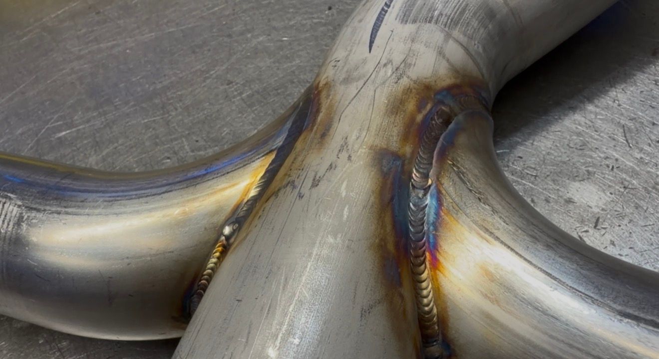 Stainless Y-pipe merge on a steel bench, an even stack-of-dimes TIG bead with gold-to-blue heat tint.